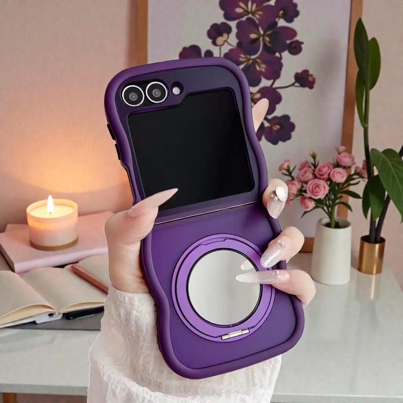 May include: A purple phone case with a built-in mirror and camera cutouts. The case is open, revealing the phone's screen. The case has a smooth, matte finish and a circular mirror on the bottom half. The phone case is designed to protect the phone.