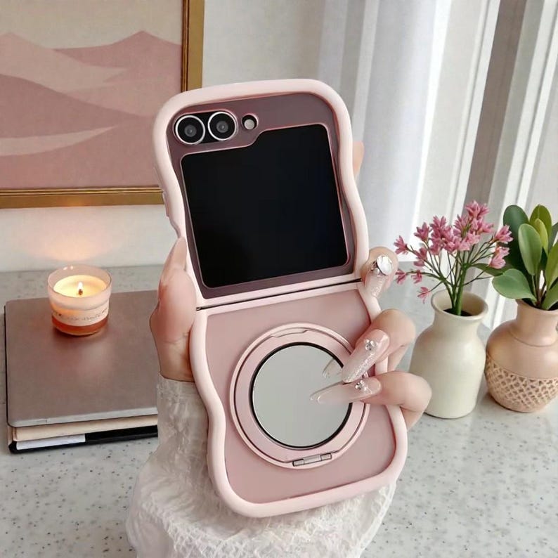 May include: A pink phone case with a built-in mirror and camera cutouts. The case is open, revealing a black screen. The design includes a rounded shape and a decorative ring. The case is held in a hand with long, decorated nails.