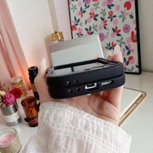 May include: A black phone case with a square design is held in a hand. The case has a raised edge around the screen and cutouts for the charging port and buttons. The case is made of a matte material.