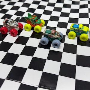 May include: Four toy monster trucks with oversized wheels are arranged on a black and white checkered surface. The trucks are various colors, including white with red wheels, green with yellow wheels, green and brown with gray wheels, and yellow with blue accents and yellow wheels.