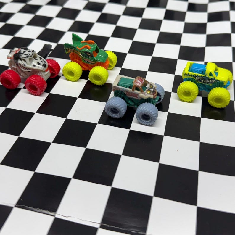 May include: Four toy monster trucks with oversized wheels are arranged on a black and white checkered surface. The trucks are various colors, including white with red wheels, green with yellow wheels, green and brown with gray wheels, and yellow with blue accents and yellow wheels.
