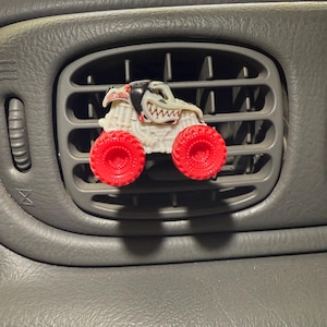 May include: A white and red monster truck air freshener is clipped into a gray car vent. The truck has red wheels and a detailed, cartoonish design. The car's dashboard is gray.