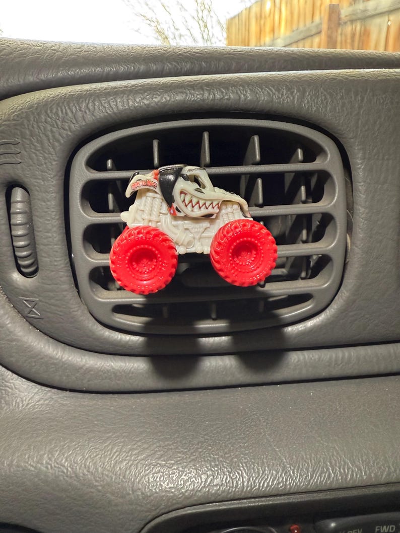 May include: A white and red monster truck air freshener is clipped into a gray car vent. The truck has red wheels and a detailed, cartoonish design. The car's dashboard is gray.