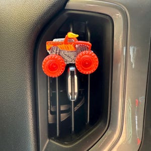 May include: A red and orange monster truck air freshener with a yellow duck on top. The truck has large orange wheels and a flame design. The text "El Monster Loco" is visible on the side.