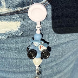 May include: A light blue retractable badge reel with a white disc, a blue bead, and a small monster truck charm. The monster truck is light blue with black wheels and a red and white design.