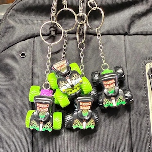 May include: Three Grave Digger monster truck keychains featuring black bodies with green flame graphics, silver accents, and bright green wheels. Each miniature truck is attached to a silver metal split ring by a short silver chain.