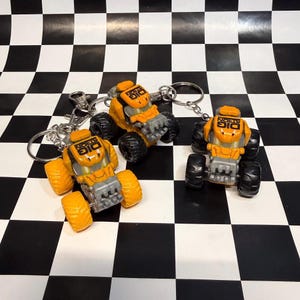 May include: Three yellow and gray monster truck keychains with black tires. Each truck features the text DIG TRON printed in black on the roof. The keychains include silver metal rings and clips for attachment, showcasing a rugged construction.
