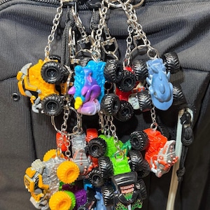 May include: A collection of monster truck keychains in various colors, including yellow, red, green, and blue. Each keychain features a miniature monster truck with large tires and a metal chain and ring for attachment.