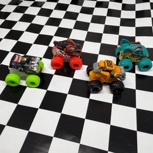 May include: Four toy monster trucks in various colors are arranged on a black and white checkered surface. The trucks include a green and black truck with green wheels, a red and black truck with red wheels, a yellow truck with black wheels, and a teal truck with teal wheels.