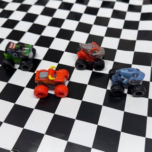May include: Four miniature monster truck toys in various colors, including green, red, and blue, with black wheels. The trucks are arranged on a black and white checkered surface. The trucks have different designs.