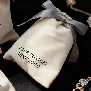 May include: Three beige velvet drawstring bags with satin ribbons in coral, grey, and peach. Each bag has the text "YOUR CUSTOM TEXT/ LOGO" printed in black. The bags are on a black surface, with jewellery in the background.