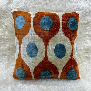 Può includere: Cuscino quadrato decorativo con un motivo ripetuto di forme a goccia arancioni e verde acqua su sfondo crema. Il cuscino è realizzato in un tessuto morbido e strutturato ed è posto su una superficie bianca e soffice.