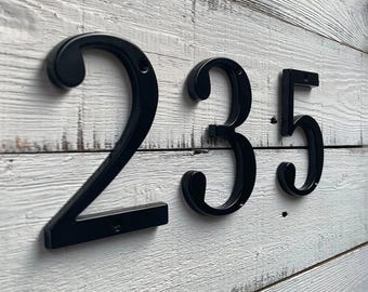May include: Black metal house numbers 235 mounted on a white-painted wooden surface. The numbers are in a classic serif font, with a slightly curved design. The wood has a distressed, rustic appearance.