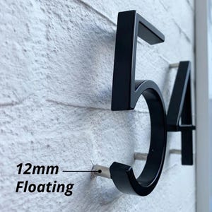 May include: Black metal house numbers, displaying "54", mounted on a white brick wall. The numbers are in a modern font and appear to float slightly off the wall. The text "12mm Floating" is also visible.