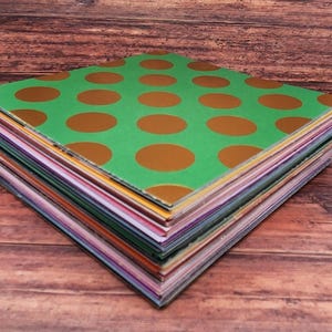 May include: A stack of colorful patterned paper sheets. The top sheet is green with large, gold-colored polka dots. The edges of the paper reveal a variety of colors, including pink, purple, yellow, and blue. The paper is stacked on a wooden surface.