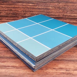 May include: A stack of square tiles with a blue color scheme. The tiles are arranged in a grid pattern, with varying shades of blue and white lines separating the squares. The tiles are stacked on a wooden surface.