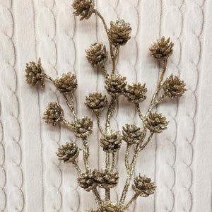 May include: A decorative floral stem with multiple golden pinecone-shaped ornaments. The ornaments and stems are covered in glitter, adding a sparkling effect. The background is a white cable-knit fabric.