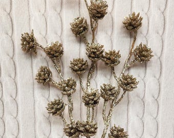 May include: A decorative floral stem with multiple golden pinecone-shaped ornaments. The ornaments and stems are covered in glitter, adding a sparkling effect. The background is a white cable-knit fabric.