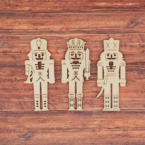 May include: Three wooden nutcracker figurines with detailed cut-out designs. Each figure has a unique hat, facial features, and accessories, including a horn, sword, and saber. The figurines are displayed on a wooden surface.