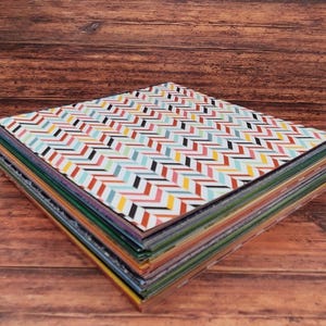 May include: A stack of colorful patterned paper sheets. The top sheet features a repeating chevron design in red, black, yellow, and blue on a white background. The edges of the paper reveal a variety of colors, including green, orange, and purple.