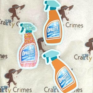 May include: Three orange spray bottle stickers with light blue spray nozzles. Each sticker has the word "OPALITE" in a blue and white design, with the text "MAGICAL RESULTS! DON'T SWEAT IT!" The background is a white surface with "Crafty Crimes" text.