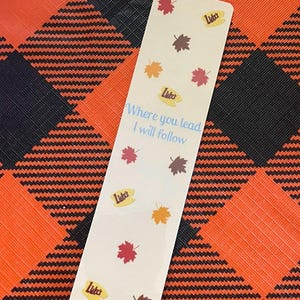 May include: A rectangular bookmark with a cream background, featuring colorful autumn leaves and the text "Where you lead I will follow." The bookmark also includes the text "Luke's" in a yellow oval. The background is a black and orange plaid pattern.