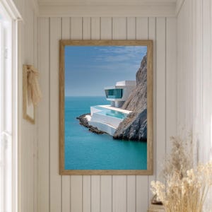 May include: A framed print of a modern white building with a pool, built into a cliffside overlooking turquoise water. The print hangs on a white paneled wall in a hallway with a woven rug.