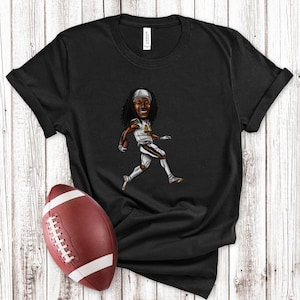 May include: Black t-shirt featuring a cartoon illustration of a football player in a white and gold uniform. A brown football is positioned near the shirt. The shirt is on a white wooden surface.