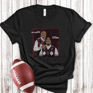 May include: Black t-shirt featuring a graphic of two men in argyle vests, with the names Terry McLaurin and Jayden Daniel. A maroon and white football is in the foreground, set against a wooden background.