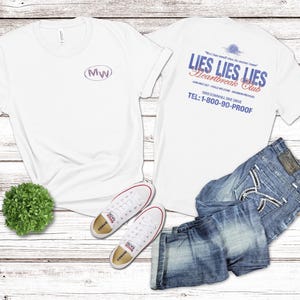 May include: White t-shirts with text and a pair of blue jeans and white sneakers. One shirt has the letters "MW" inside an oval. The other shirt has the text "LIES LIES LIES Heartbreak Club" and a phone number.