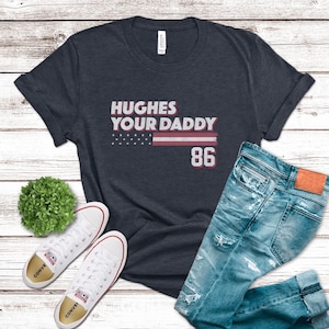 Puede incluir: Una camiseta azul marino con el texto "HUGHES YOUR DADDY" sobre un dise&ntilde;o de bandera estadounidense y el n&uacute;mero "86". La camiseta se combina con vaqueros azules desgastados y zapatillas blancas.