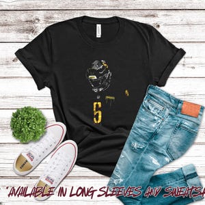 May include: Black t-shirt with a football helmet graphic and the number 5 in yellow. The shirt is paired with blue jeans and white Converse sneakers. A small green plant is also in the image.