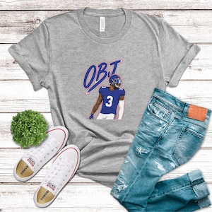 May include: A heather gray t-shirt featuring a graphic of a football player wearing a blue jersey with the number 3 and the text OBJ in blue and red. The shirt is displayed alongside a pair of distressed blue jeans and white canvas sneakers.