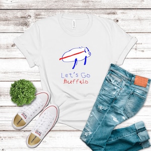 May include: White t-shirt with a blue and red cartoon buffalo and the text "Let's Go Buffalo." The shirt is paired with blue jeans and white sneakers with red accents. The items are arranged on a light wood-grain surface.