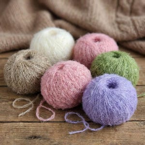 以下が含まれることがあります： 白、ピンク、緑、紫、茶色の色合いのカラフルな毛糸玉のコレクション。 毛糸玉は木製の表面に配置され、背景には柔らかく質感のある生地があります。 糸はふわふわで柔らかく見えます。