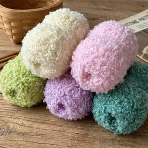 May include: Six balls of colourful yarn in shades of green, cream, pink, purple, and teal. The yarn has a textured, looped appearance, suggesting a soft, plush material. The balls are arranged on a wooden surface.
