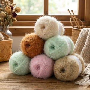 May include: A stack of fluffy yarn balls in various pastel colors, including cream, light green, pink, and brown. The yarn is arranged on a wooden surface, suggesting a craft or knitting project. A basket and knitting needles are in the background.