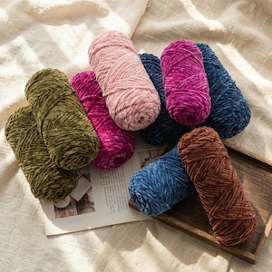 May include: A collection of colorful velvet yarn skeins in shades of olive green, pink, fuchsia, navy blue, light blue, and brown. The yarn is arranged on a wooden tray and a newspaper, set against a cream-colored fabric background.