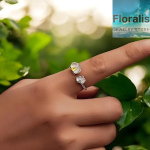 May include: A silver ring with two flower designs, one on top and one below. The flowers have white petals and yellow centers. The ring is worn on a finger. The image also includes the text "Floralis JEWELRY STORE".