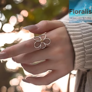 Puede incluir: Un anillo de flores plateado con un centro dorado se muestra en un dedo. El anillo presenta cinco delicados pétalos. La imagen también muestra el texto "Floralis Jewelry Store".