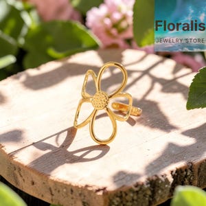 May include: A gold-colored flower ring with a detailed center and open petals. The ring is displayed on a wooden surface with the text "Floralis Jewelry Store" in the background. The ring's design is floral and elegant.