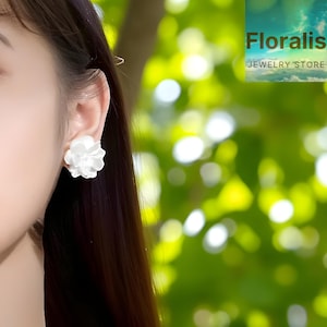 Può includere: Primo piano di un orecchino bianco a forma di fiore. L'orecchino è indossato su un orecchio, con il logo Floral Jewelry Store sullo sfondo. Lo sfondo è un verde sfocato.
