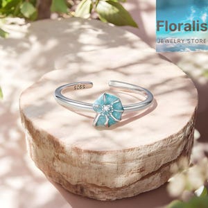 May include: A silver adjustable ring with a light blue flower design and a small, clear gemstone. The ring is marked "S925". The Floralist Jewelry Store logo is visible in the background.