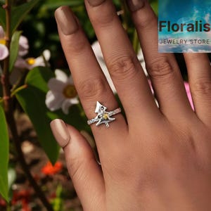 Puede incluir: Un anillo plateado con un diseño de árbol de Navidad, con una piedra preciosa amarilla y pequeñas piedras blancas. El anillo está en una mano con un fondo borroso de follaje verde. El texto "Floralis Jewelry Store".
