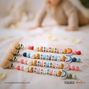 Peut inclure: Trois attaches-sucettes pour bébés avec des perles en bois et en silicone. Chaque attache affiche "BABY" avec des perles de lettres et présente un motif arc-en-ciel. Les attaches ont des attaches en bois et sont de couleurs pastel variées.