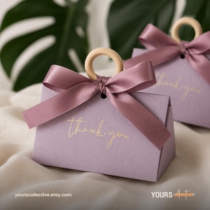 May include: Two light purple gift boxes with embossed floral patterns and gold foil text that reads thank you. Each box features a satin mauve ribbon tied in a bow around a circular wooden handle, creating a decorative closure for the container.