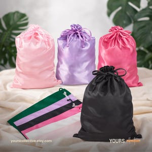 May include: A collection of satin drawstring bags in various colors: pink, purple, and black. A flat, multi-colored bag with green, black, pink, and white stripes is also present. The bags are arranged on a neutral-toned surface. The website yours collective etsy com is visible.