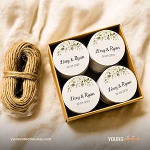May include: A box of round white stickers with floral designs and the text "Lizzy & Ryan 20.09.2025". A bundle of twine sits next to the box. The stickers are in a gold box. The background is a light beige fabric.