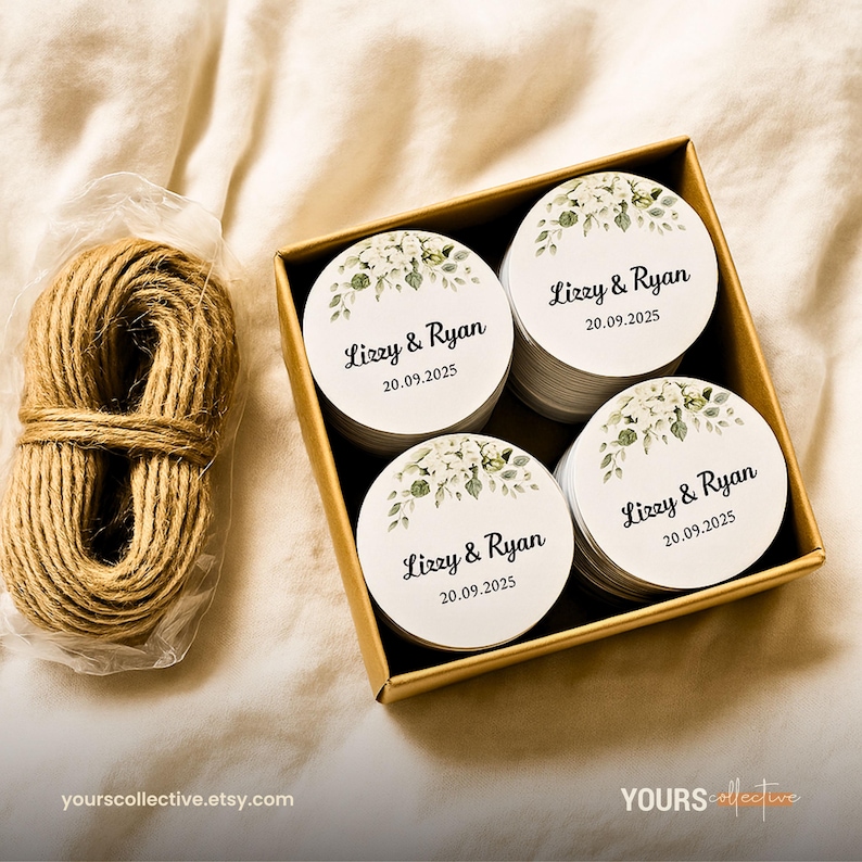 May include: A box of round white stickers with floral designs and the text "Lizzy & Ryan 20.09.2025". A bundle of twine sits next to the box. The stickers are in a gold box. The background is a light beige fabric.
