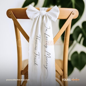 Puede incluir: Una silla de madera con un lazo y una faja de tela color crema. La faja tiene el texto "In Loving Memory Thomas Frank" en escritura negra. La silla es de madera clara.